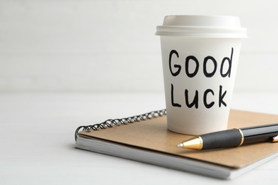 Photo of Paper coffee cup with phrase GOOD LUCK and stationery on white wooden table, closeup. Space for text