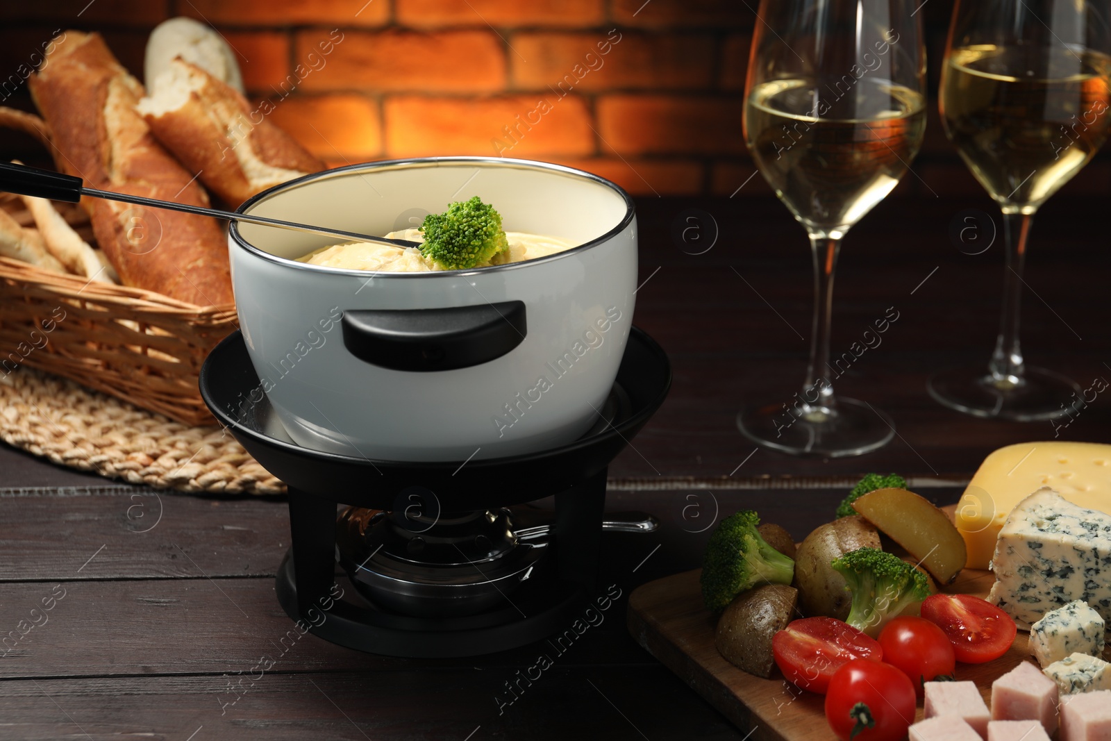 Photo of Fork with piece of broccoli, melted cheese in fondue pot, wine and snacks on wooden table, closeup