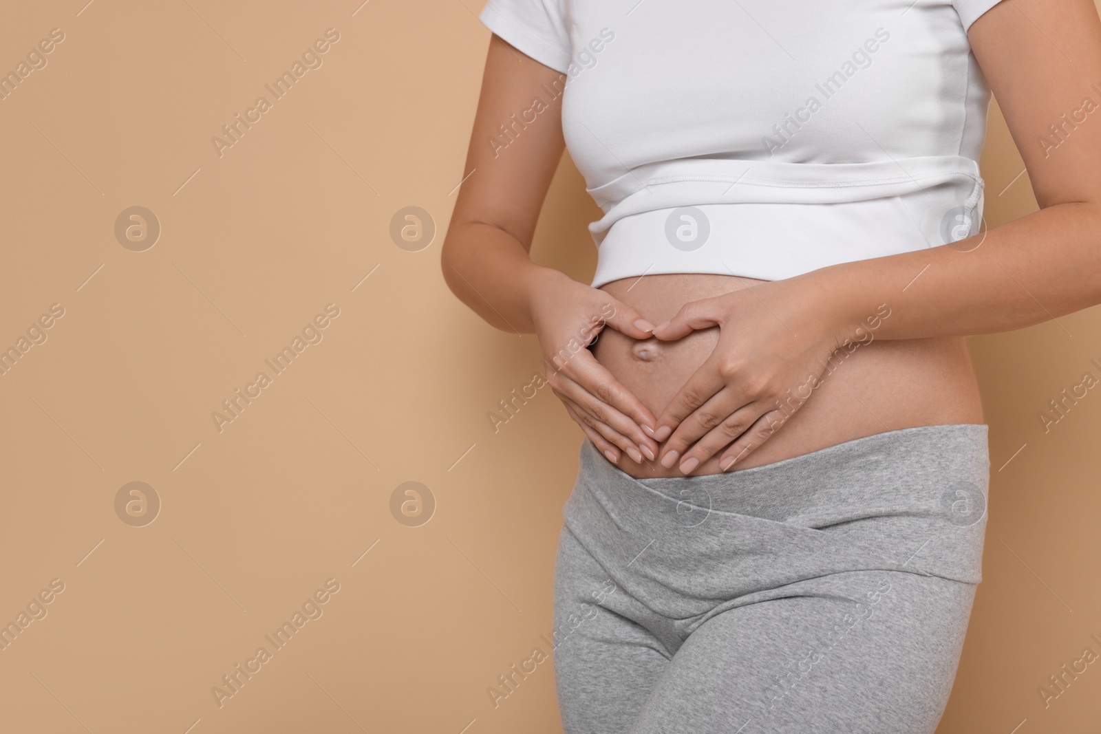 Photo of Pregnant woman making heart with hands on beige background, closeup. Space for text