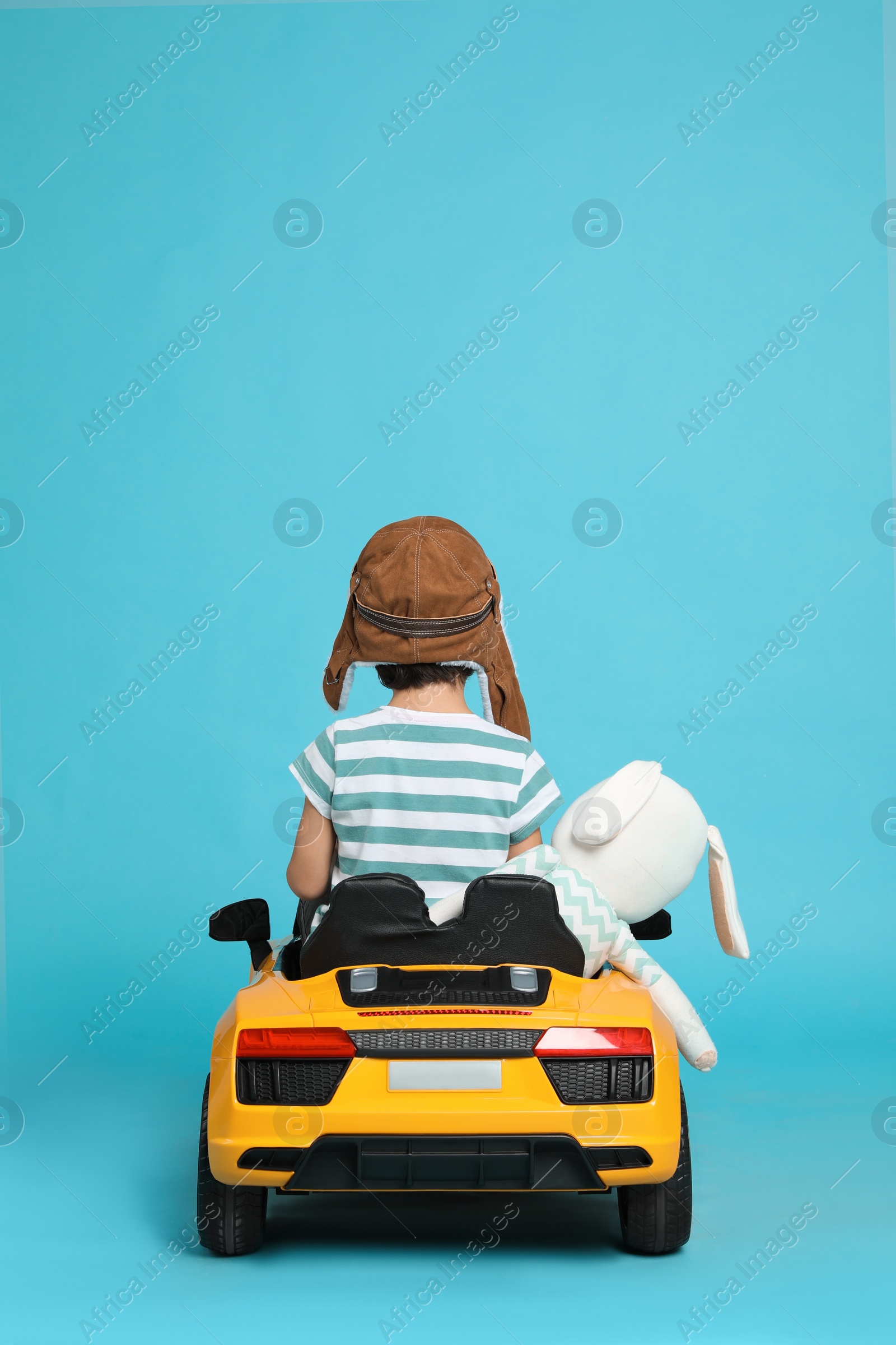 Photo of Cute little boy with toy bunny driving children's car on light blue background, back view