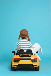 Photo of Cute little boy with toy bunny driving children's car on light blue background, back view