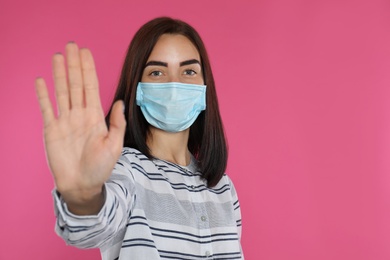 Photo of Young woman in protective mask showing stop gesture on pink background, space for text. Prevent spreading of coronavirus
