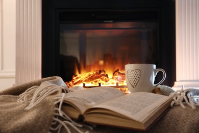 Photo of Cup of hot drink and book on blanket near fireplace indoors. Magic atmosphere