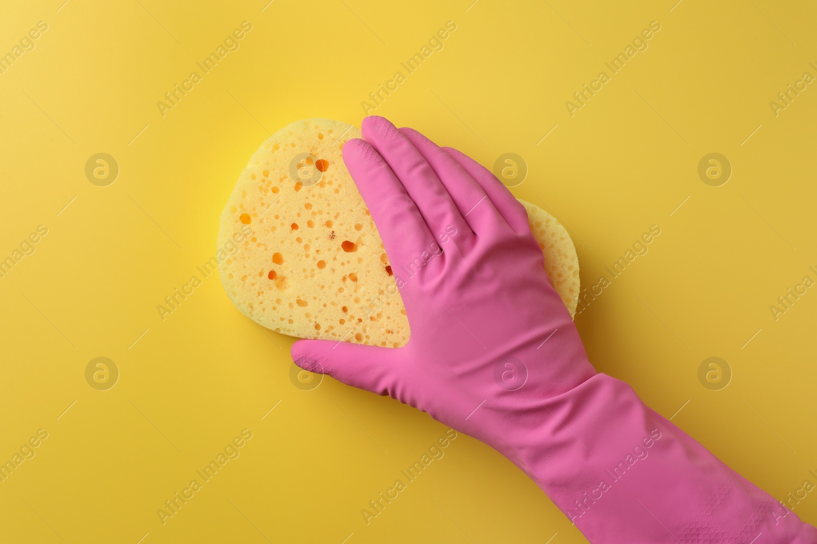 Photo of Cleaner in rubber glove holding new sponge on yellow background, top view