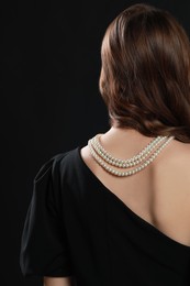 Young woman wearing elegant pearl necklace on black background, back view