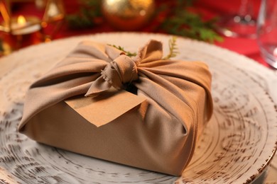 Photo of Furoshiki technique. Gift packed in beige fabric, blank card and festive decor on plate, closeup