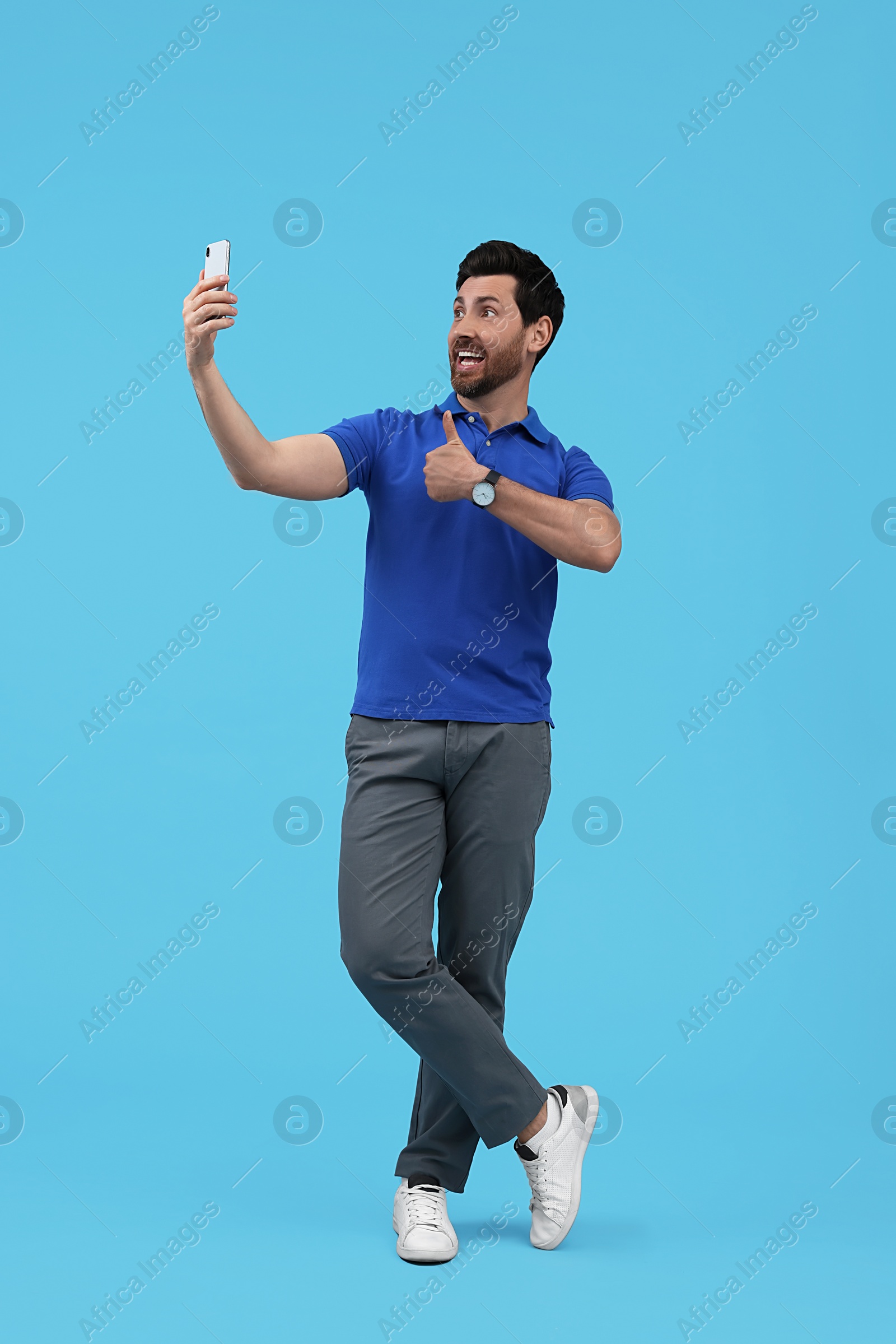 Photo of Smiling man taking selfie with smartphone and showing thumbs up on light blue background