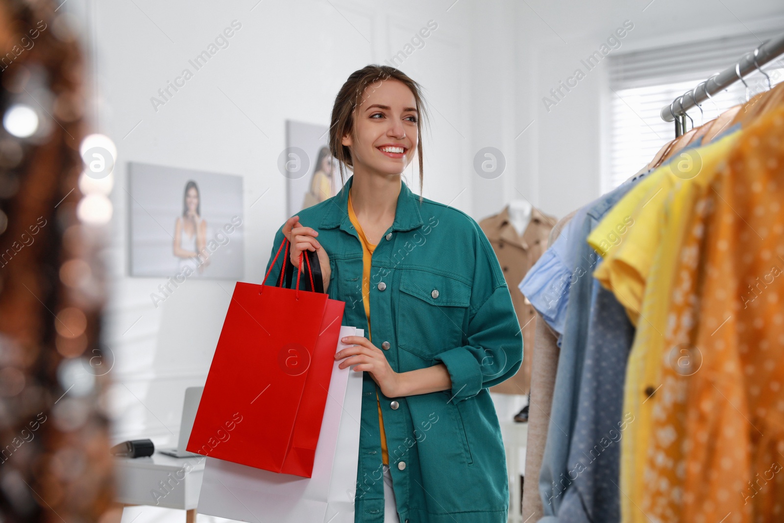 Photo of Young woman holding shopping bags near rack with clothes in modern boutique