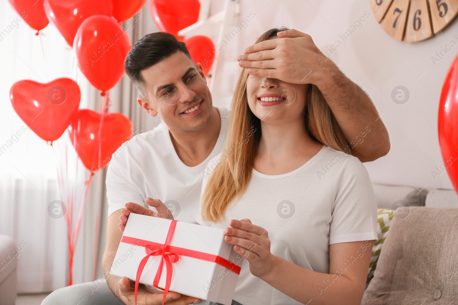 Photo of Man presenting gift to his girlfriend in room decorated with heart shaped balloons. Valentine's day celebration