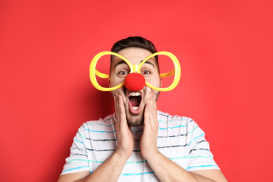 Photo of Emotional young man with party glasses and clown nose on red background. April fool's day