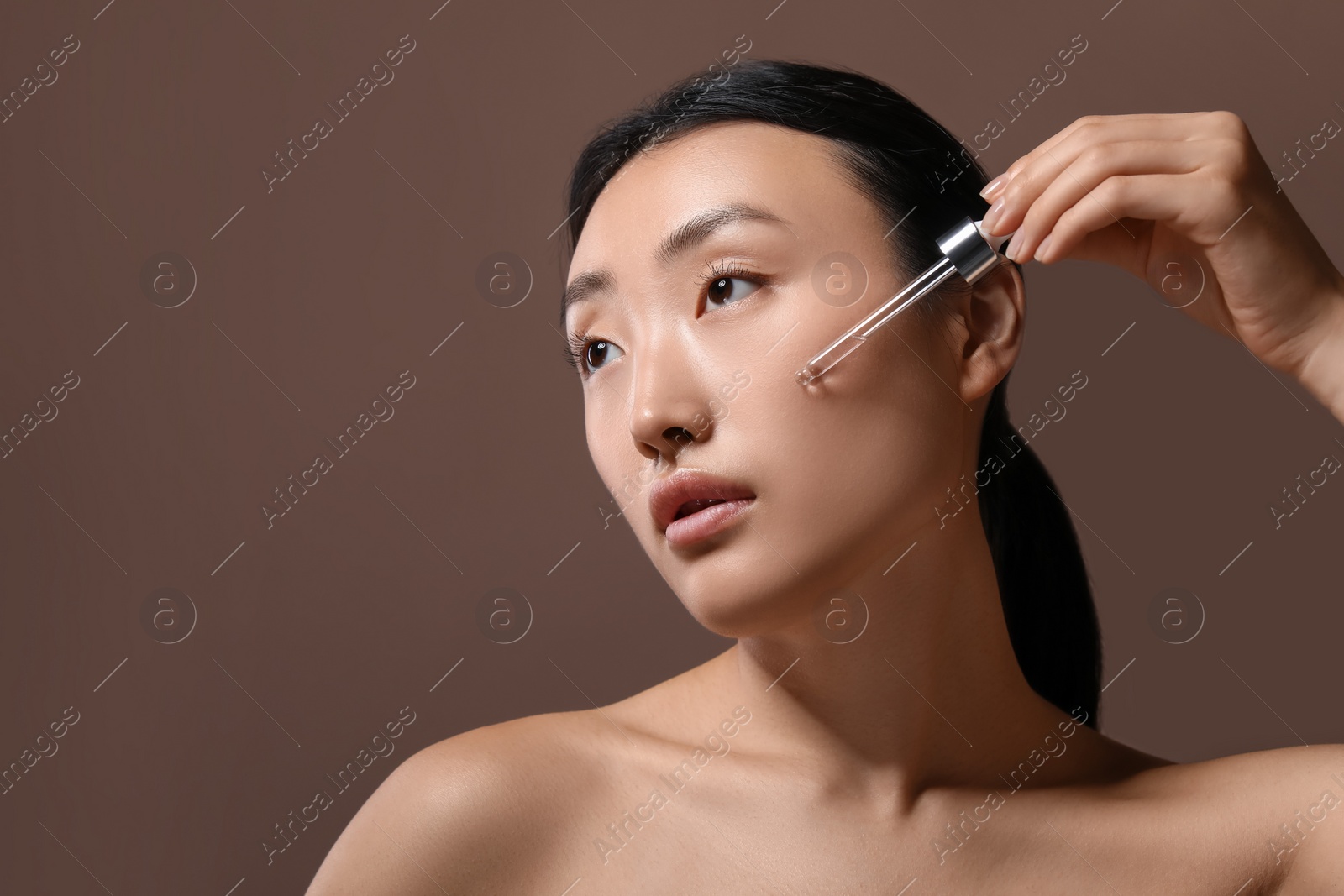 Photo of Beautiful young woman applying cosmetic serum onto her face on brown background, space for text