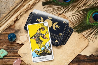 Photo of The Fool near other tarot cards with peacock feathers, gemstones and old book on wooden table, above view