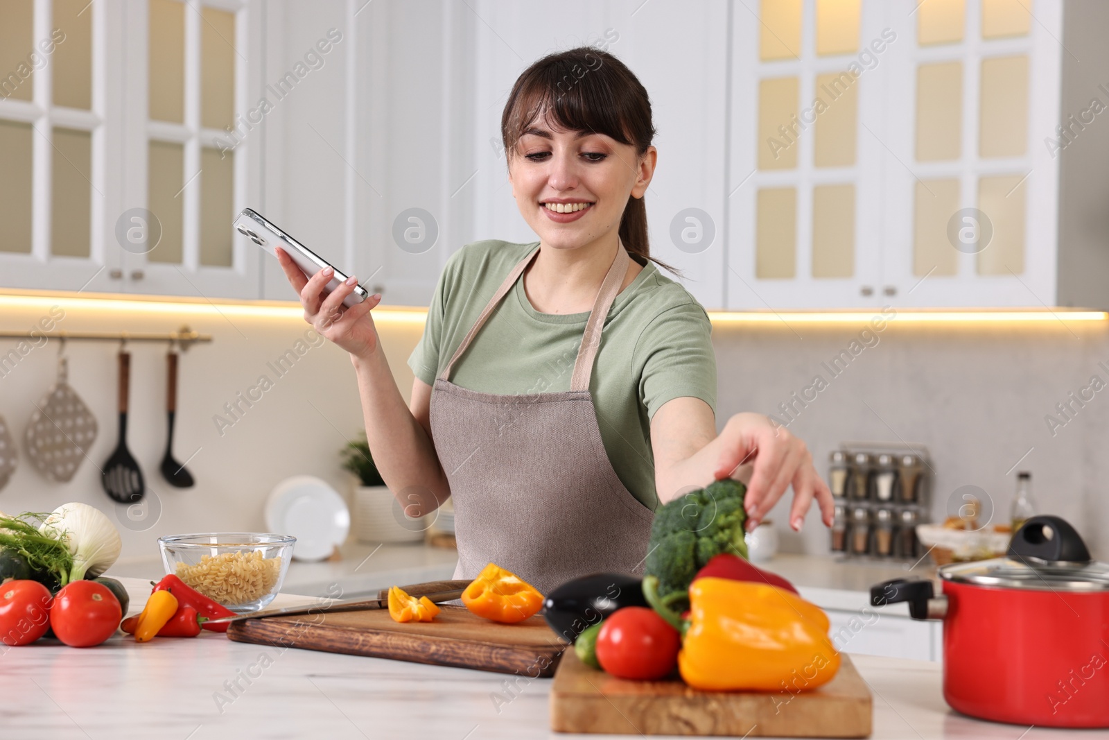 Photo of Happy young housewife using smartphone while cooking at white marble table in kitchen