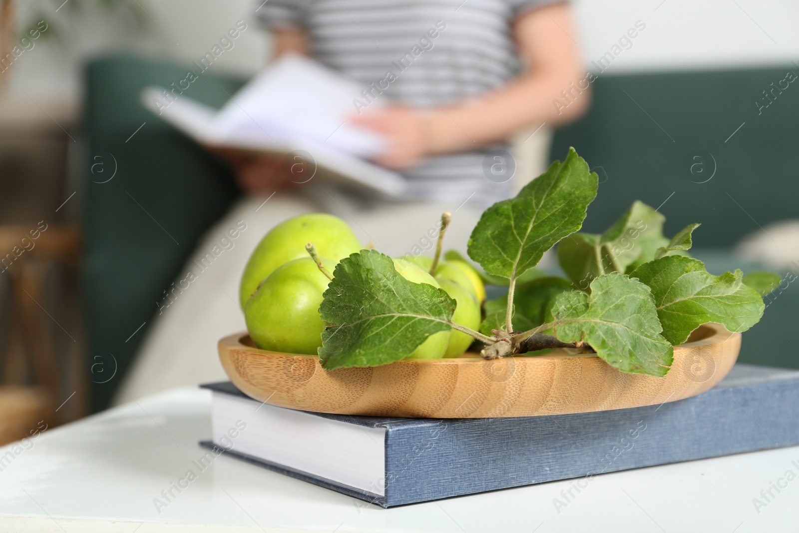 Photo of Woman reading book in living room, focus on green apples