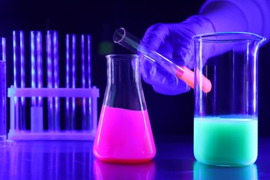 Photo of Scientist pouring luminous liquid into laboratory glassware at table, selective focus