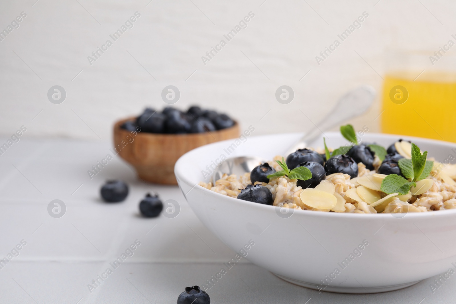 Photo of Tasty oatmeal with blueberries, mint and almond flakes in bowl on white tiled table, closeup. Space for text