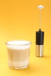 Photo of Mini mixer (milk frother) and tasty cappuccino in glass on yellow background