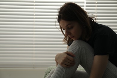 Sad young woman near window indoors, space for text