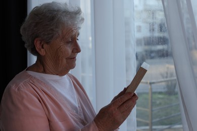Photo of Elderly woman with photo frame near window indoors. Loneliness concept