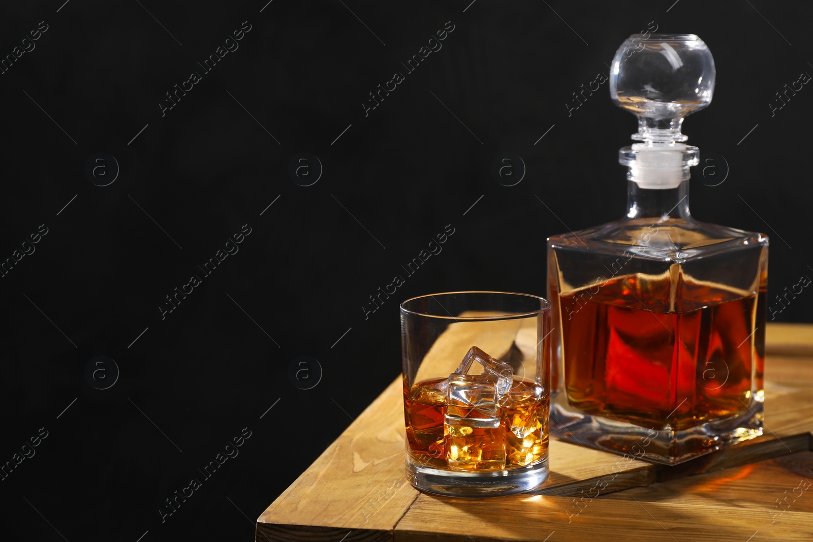 Photo of Whiskey with ice cubes in glass and bottle on wooden crate against black background, space for text