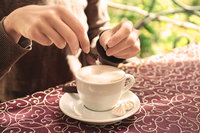 Photo of Young woman adding sugar to delicious coffee at table