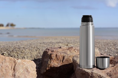 Metallic thermos and cap on stone near sea, space for text