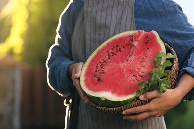 Photo of Woman holding half of delicious ripe watermelon in wicker basket with mint outdoors, closeup. Space for text