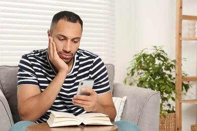 Photo of Young man using smartphone while reading book at home, space for text. Internet addiction