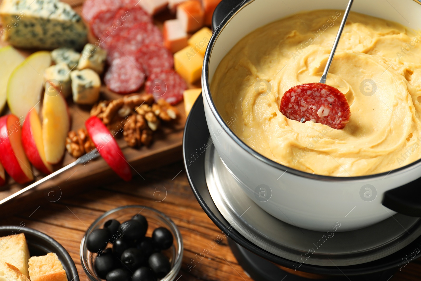 Photo of Fork with piece of sausage, melted cheese in fondue pot and other products on wooden table, closeup