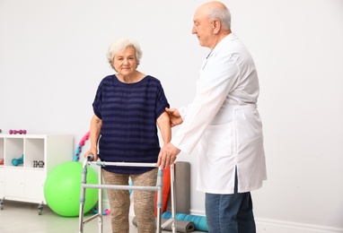 Photo of Doctor helping elderly woman with walking frame indoors