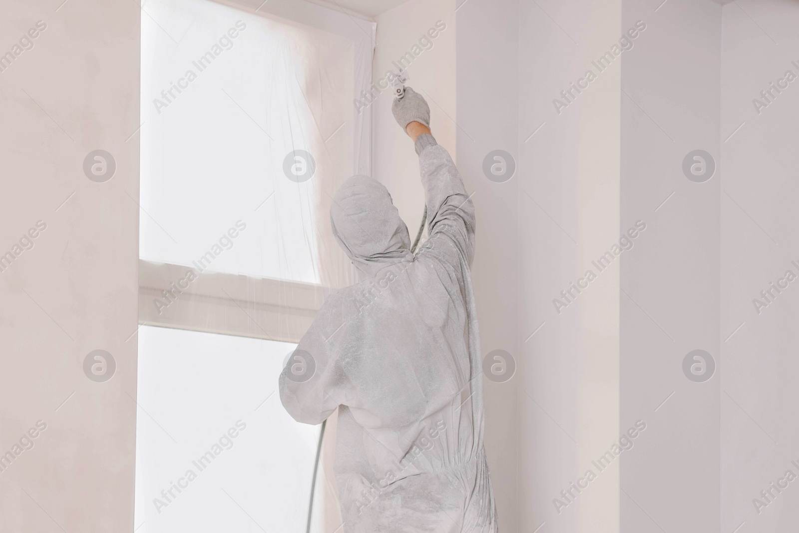 Photo of Decorator in uniform painting wall with sprayer indoors, back view