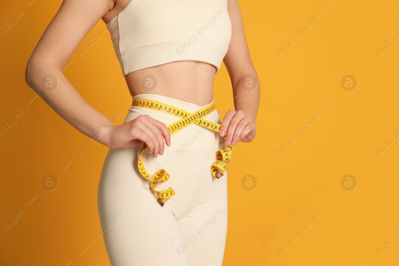 Photo of Woman in sportswear measuring waist with tape on yellow background, closeup. Space for text