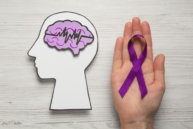 Photo of Woman holding purple ribbon near human head cutout with brain and pulse line on white wooden background, top view. Epilepsy awareness