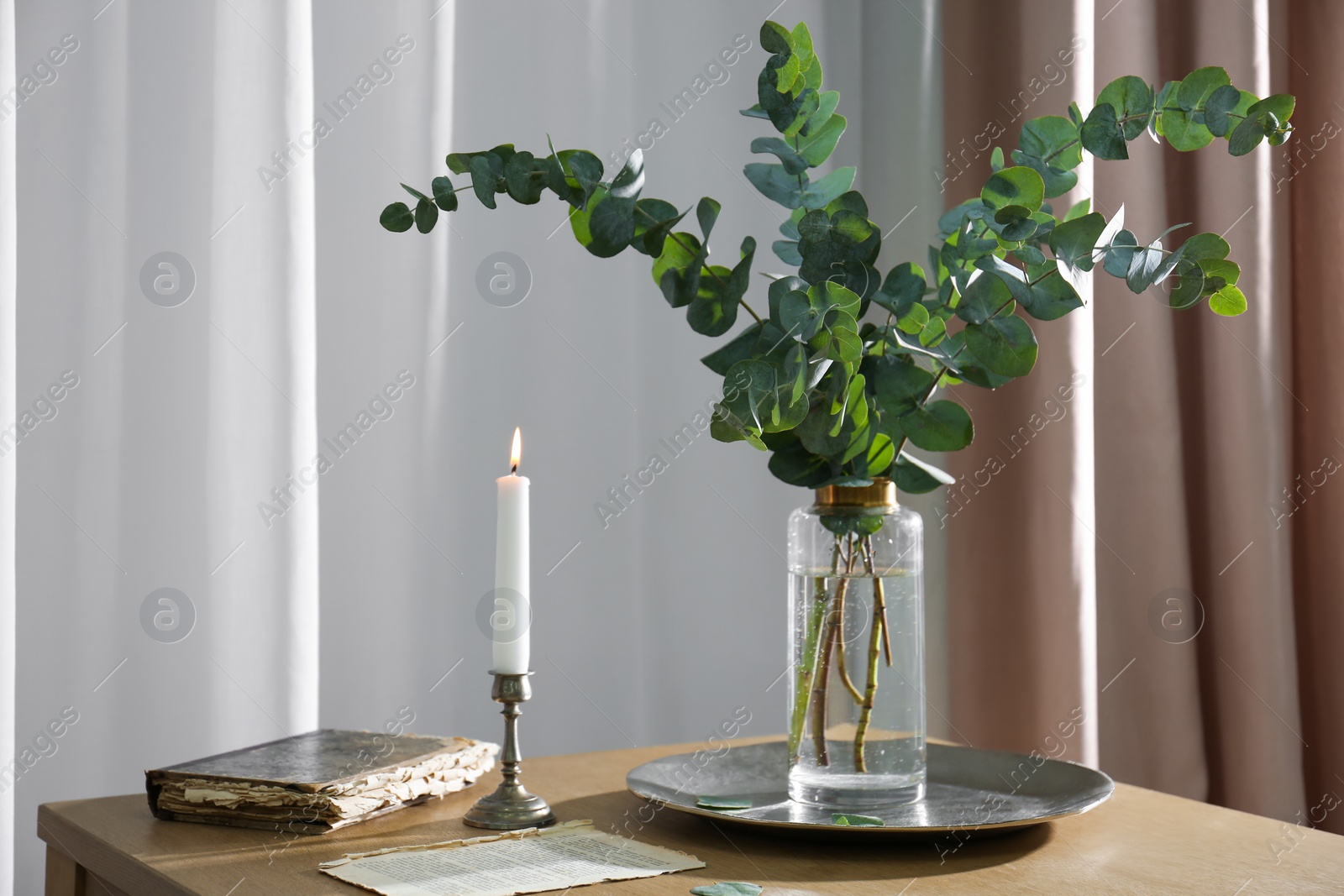 Photo of Beautiful eucalyptus branches, old book and holder with burning candle on wooden table indoors. Interior element