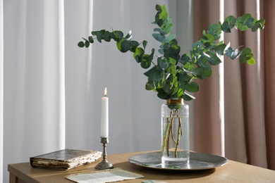 Photo of Beautiful eucalyptus branches, old book and holder with burning candle on wooden table indoors. Interior element