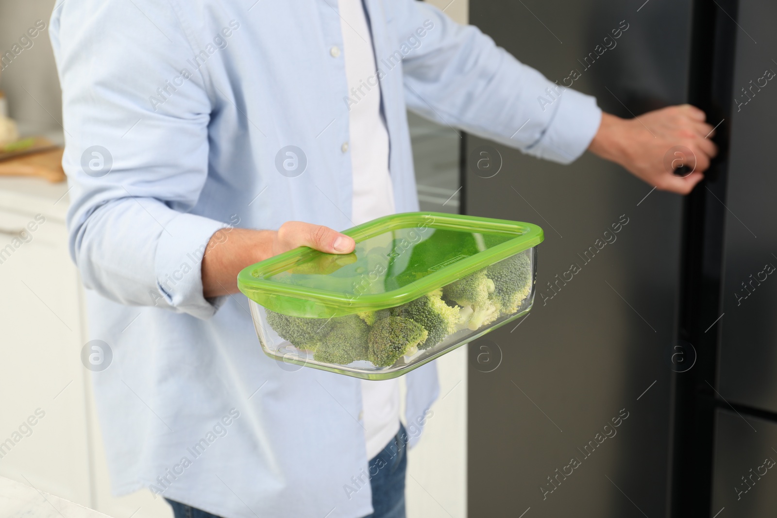 Photo of Man holding container with fresh broccoli near fridge in kitchen, closeup. Food storage