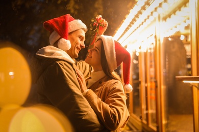 Photo of Happy couple in Santa hats standing under mistletoe bunch outdoors, bokeh effect