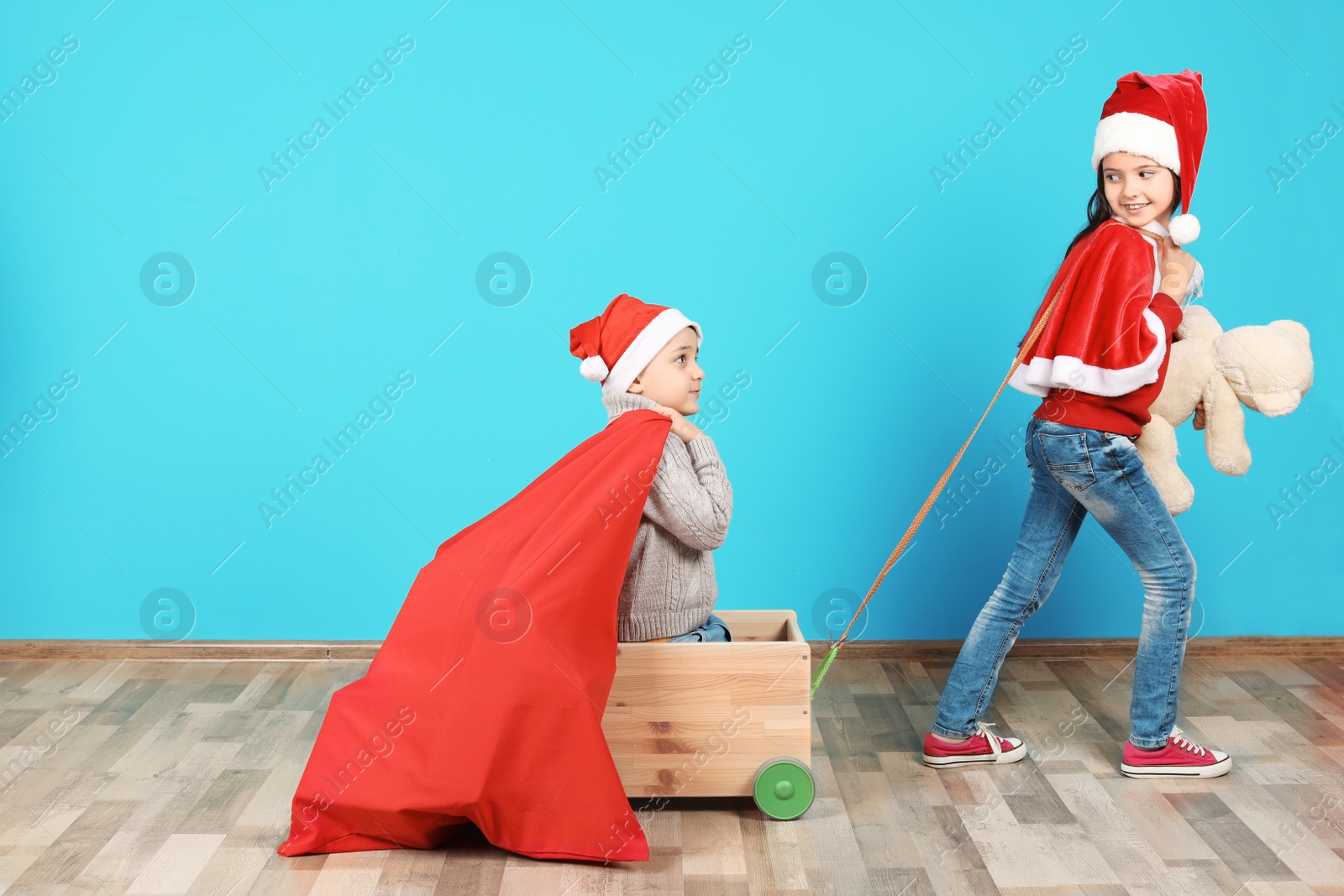 Photo of Cute little children in Santa hats playing with toy cart and Christmas sack near color wall