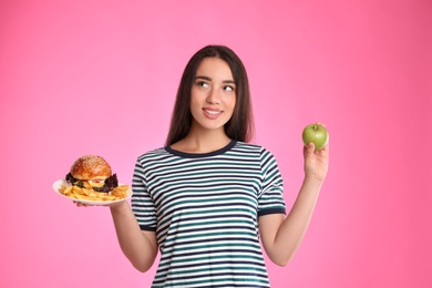 Photo of Woman choosing between apple and burger with French fries on pink background