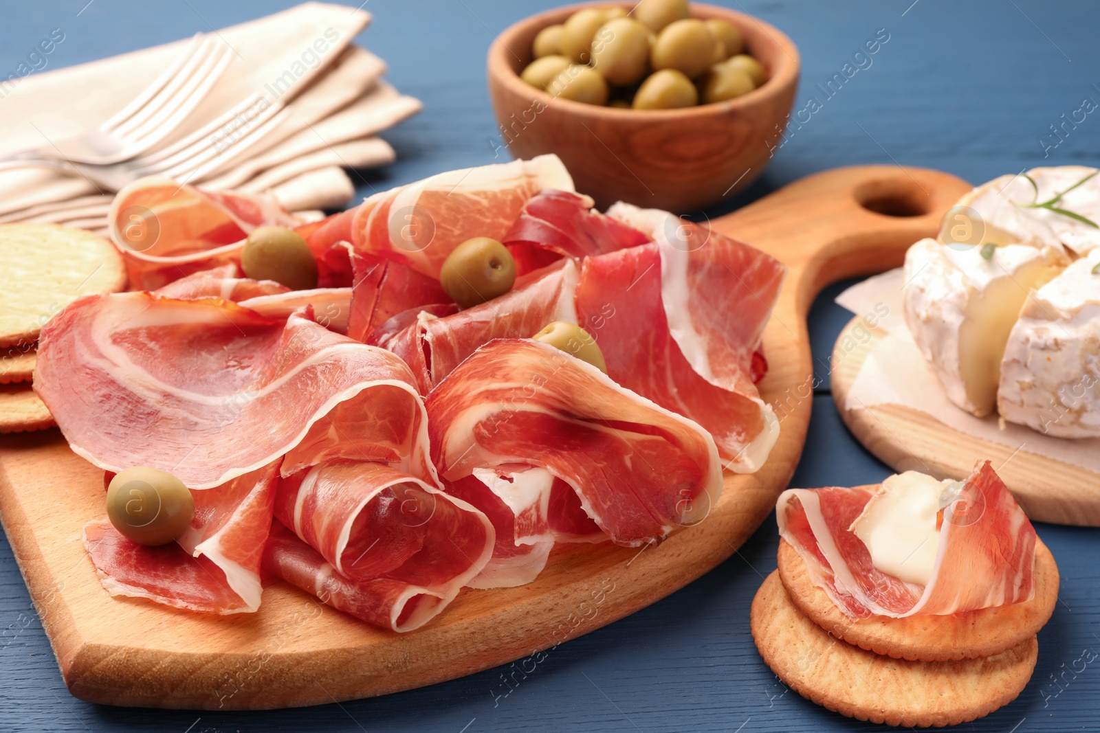Photo of Slices of tasty cured ham, olives and cheese on blue wooden table, closeup