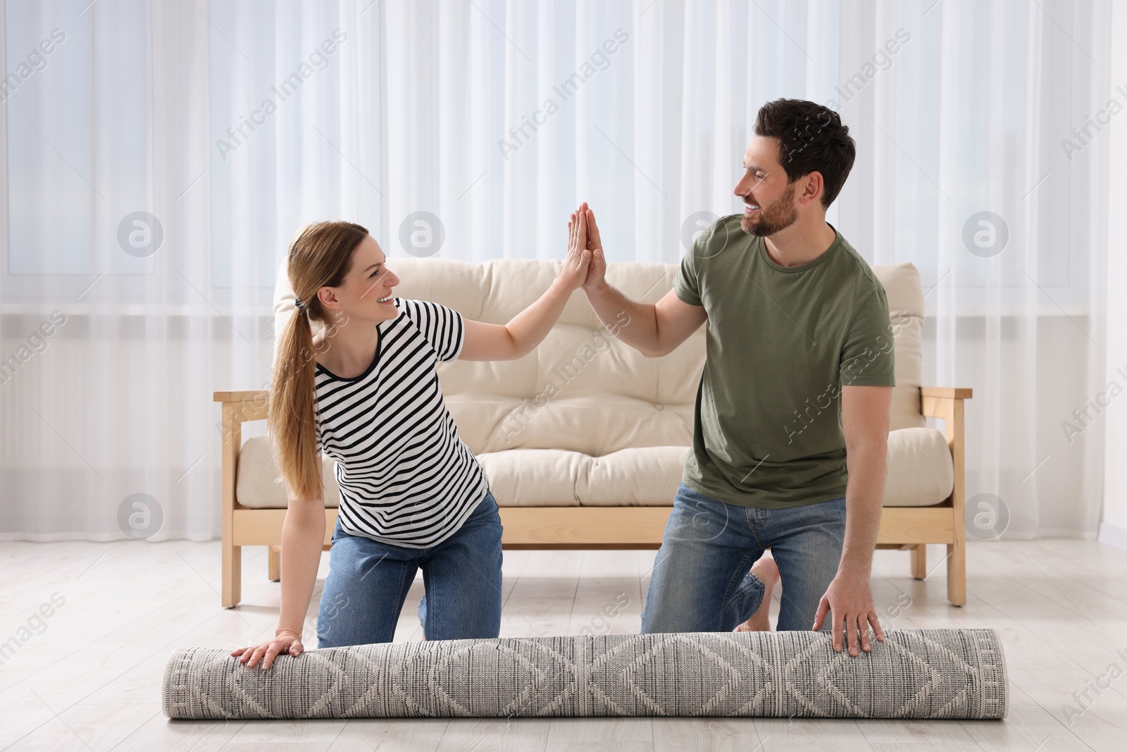 Photo of Happy couple giving each other high five while unrolling carpet on floor in room