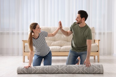 Photo of Happy couple giving each other high five while unrolling carpet on floor in room