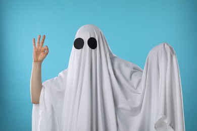 Photo of Funny ghost. Woman covered with white sheet showing OK gesture on light blue background