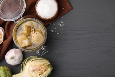 Jar of delicious artichokes pickled in olive oil and ingredients on dark grey wooden table, flat lay. Space for text