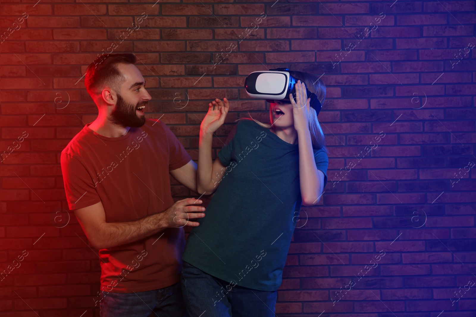 Photo of Emotional woman playing video games with VR headset and happy man near brick wall. Space for text