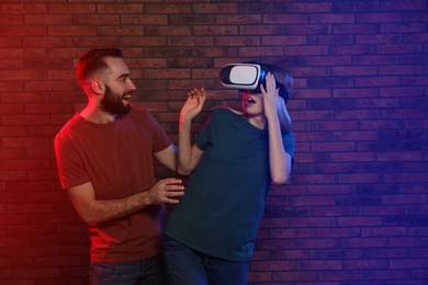 Photo of Emotional woman playing video games with VR headset and happy man near brick wall. Space for text