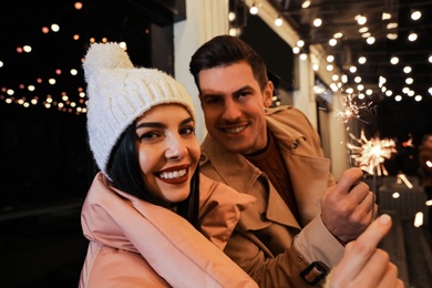 Photo of Couple in warm clothes holding burning sparklers near building