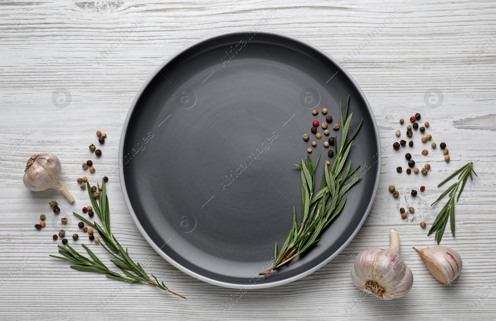 Photo of New dark plate with rosemary, garlic and peppercorns on light wooden table, flat lay