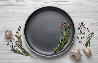 Photo of New dark plate with rosemary, garlic and peppercorns on light wooden table, flat lay