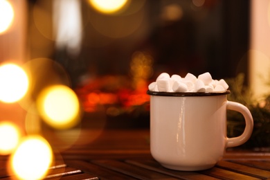 Photo of Cup of hot drink with marshmallows on wooden table near fireplace at home. Space for text